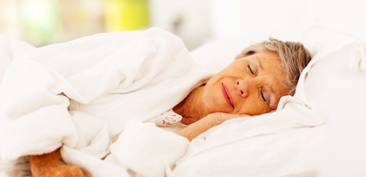 An older woman in white robe sleeping on white linen sheets.