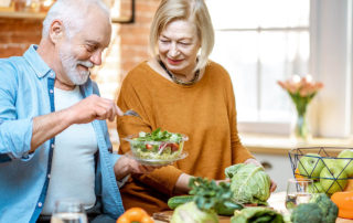 Cheerful senior couple eating salad standing together with healthy food on the kitchen at home. Concept of healthy nutrition in older age