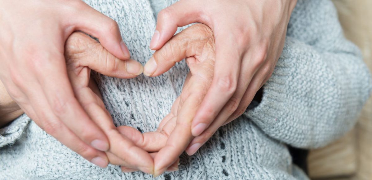 Two pairs of hands (one younger, one senior) forming a heart shape over a grey sweater.