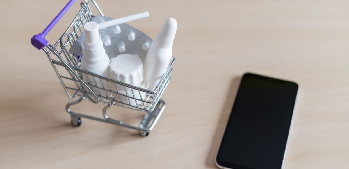 A tiny shopping cart full of medications next to a smartphone.